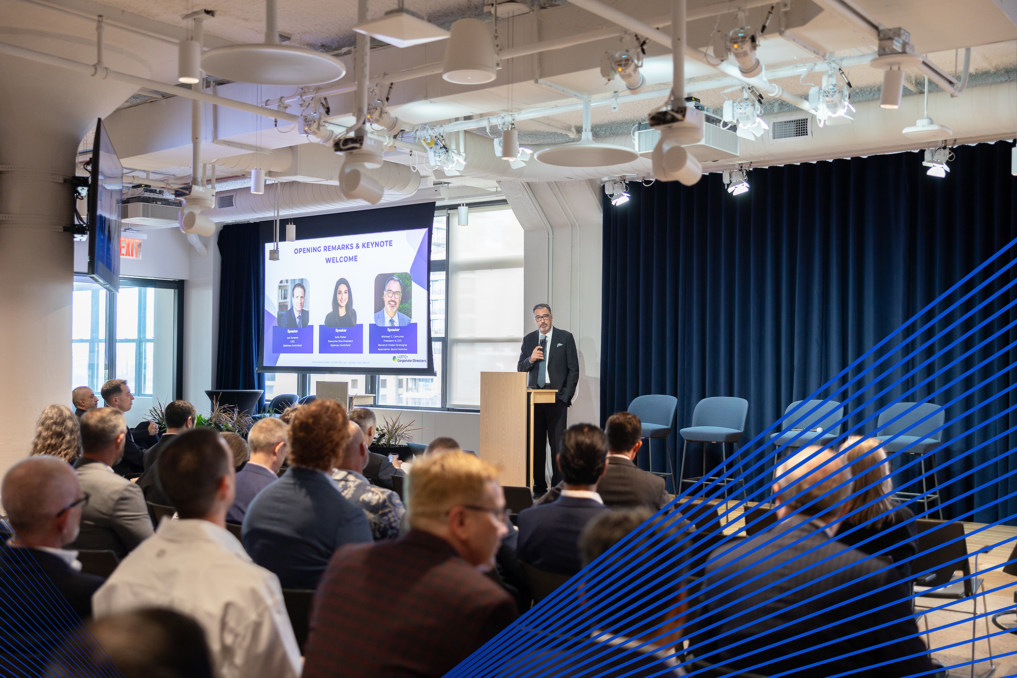 Speaker delivering opening remarks and keynote at the PRISM annual summit, with an audience of directors seated.